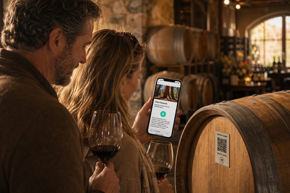 A couple in a winery barrel cellar, one holds a wine glass, the other a phone showing the Wexplo audio guide playing a stop; a numbered QR code is mounted on the barrel.